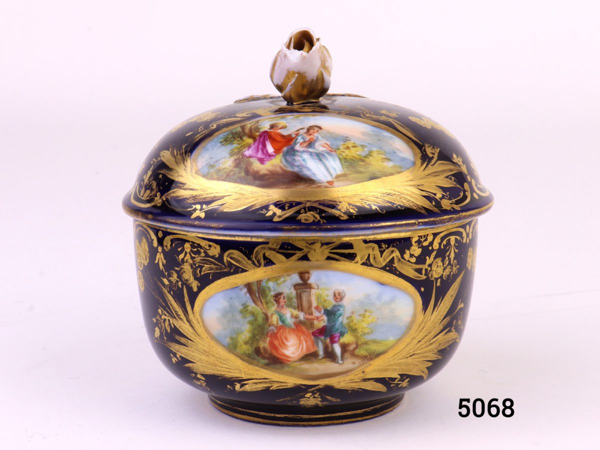 Antique Meissen small lidded bowl in royal blue with gilt throughout and decorated with intricate hand-painted images of courting couples. Rose bud to top of lid intact with minor chips at the edges and some gilt wear. Measures 45mm ín diameter at base and 80mm across the top.                               Base of lid measures 86mm Main photo of pot with lid in place showing 2 of the courting couple scenes