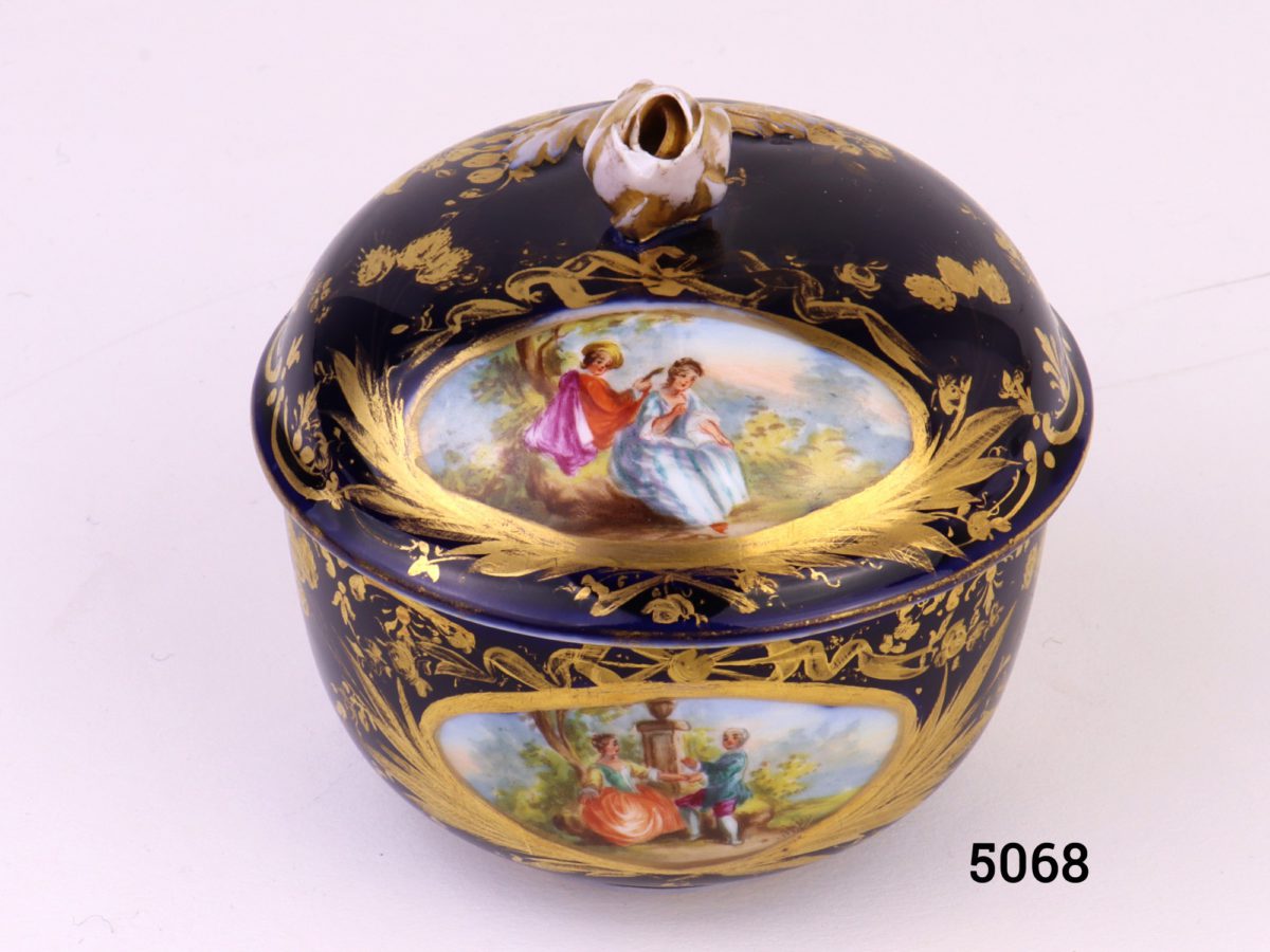 Antique Meissen small lidded bowl in royal blue with gilt throughout and decorated with intricate hand-painted images of courting couples. Rose bud to top of lid intact with minor chips at the edges and some gilt wear. Measures 45mm ín diameter at base and 80mm across the top.     Base of lid measures 86mm Photo of pot with lid in place from a slightly raised angle