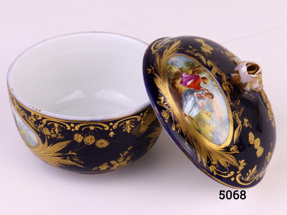 Antique Meissen small lidded bowl in royal blue with gilt throughout and decorated with intricate hand-painted images of courting couples. Rose bud to top of lid intact with minor chips at the edges and some gilt wear. Measures 45mm ín diameter at base and 80mm across the top.     Base of lid measures 86mm Photo of pot with lid removed and leaning against bottom half