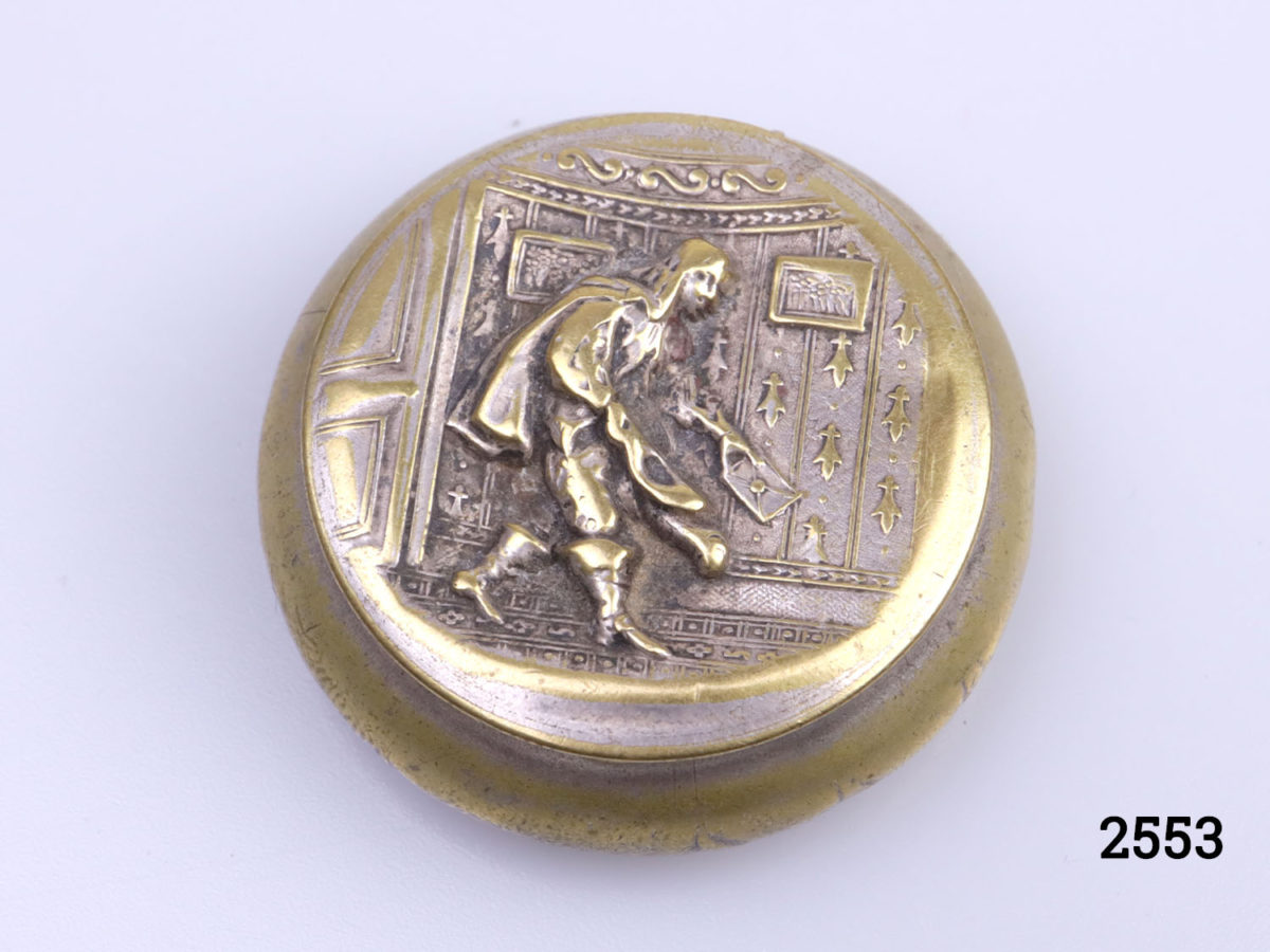Antique brass pill or snuff box. Scene of cloaked man holding a sealed letter to the lid (Possibly a Royal messenger). Foliage pattern on the reverse (base) Measures 38mm in diameter at base. Main photo of closed box showing messenger on lid