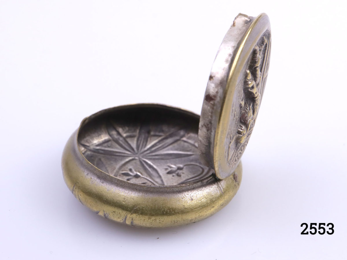 Antique brass pill or snuff box. Scene of cloaked man holding a sealed letter to the lid (Possibly a Royal messenger). Foliage pattern on the reverse (base) Measures 38mm in diameter at base. Photo of box open shown from a side angle showing relief embossing on base