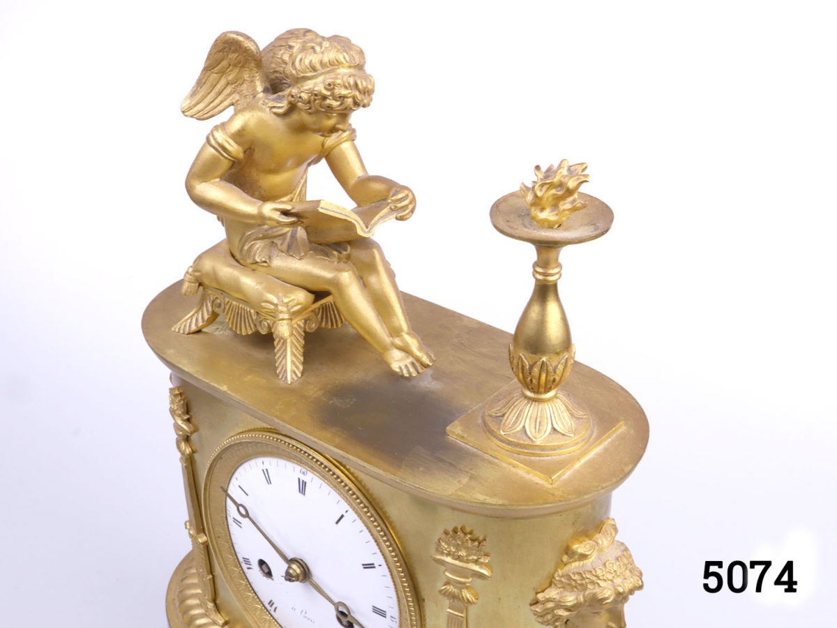 Antique French clock c1810s. Heavy clock in gilt ormolu from the first French empire. Fine detail decoration with an angel reading by firelight. Back cover missing. Key included. Fully serviced and guaranteed for 1 year. Photo of top of clock from a side angle showing detail of angel and fire column