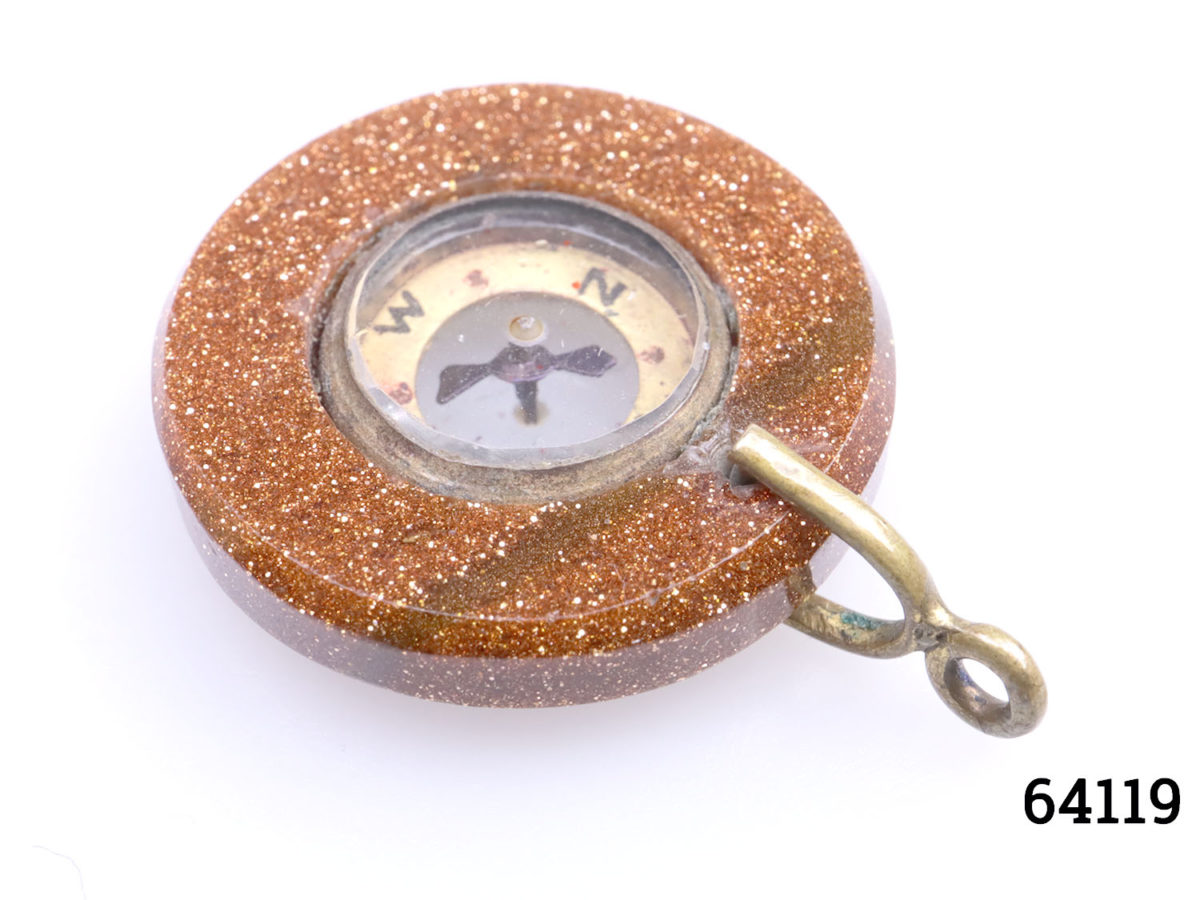 Antique goldstone compass fob. Miniature compass set in a ring of sparkly goldstone and mounted on brass. Comes in box and pouch. Measures 20mm in diameter and weighs 3.6g. Close up photo of fob with brass clasp in the foreground