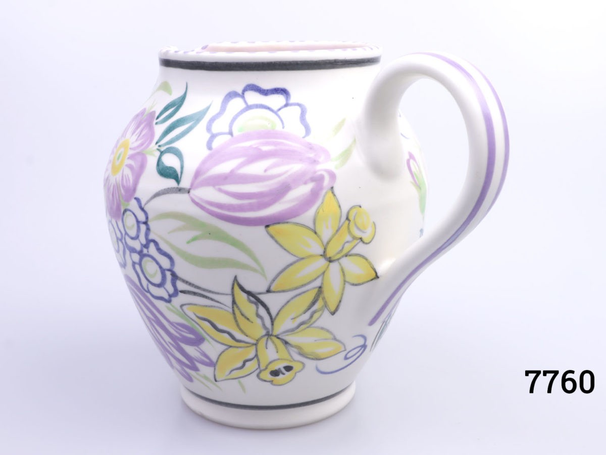 Vintage small Poole Pottery jug with spring flowers in lilac, green and yellow with pale pink interior. (Tiny chip to the lip). Measures 60mm in diameter at base. Photo of jug from an eye level angle showing the floral decoration near the handle