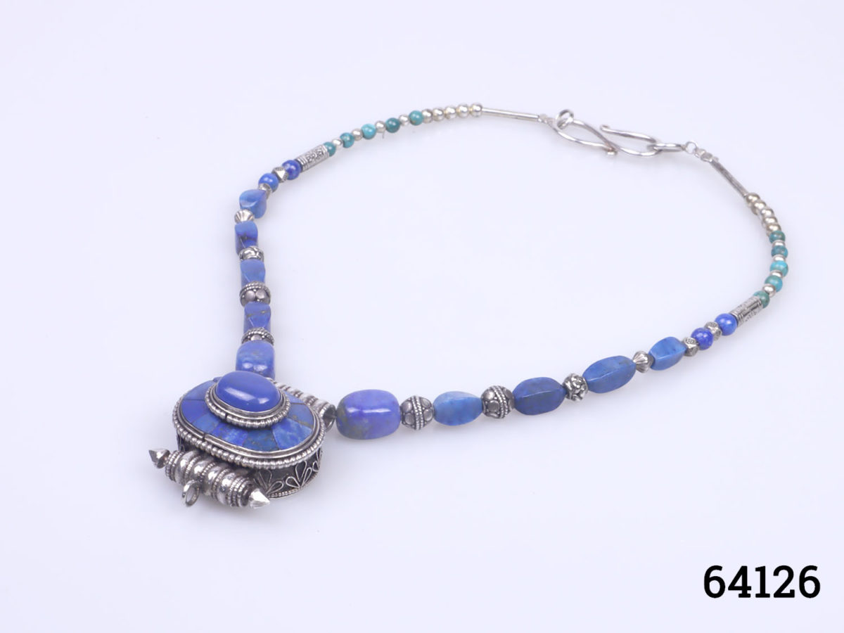 Vintage Tibetan silver necklace with turquoise and lapis lazuli beads and a lapis lazuli box pendant. Pendant has a secret compartment that opens at the back and measures 38mm long by 30mm wide Photo of necklace on a flat surface seen diagonally laid with pendant area to the bottom left