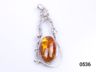 c1990s Vintage sterling silver pendant with large amber cabochon. Amber set on sterling silver in an eclectic design mix of modernist and Art Nouveau styles. Fully hallmarked for London assay. Measures 40mm at widest point. Drop length 115mm from bail to bottom of pendant. Main photo of pendant on a flat surface shown as it would be worn with amber in the bottom centre