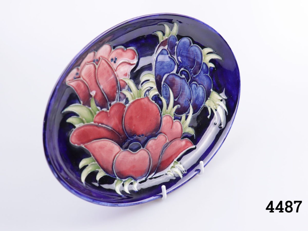 Vintage Moorcroft oval plate in cobalt blue anemone design with dark pink and blue anemone flowers. Pattern 1930. Photo of dish on a display stand from a slight side angle with dish facing right