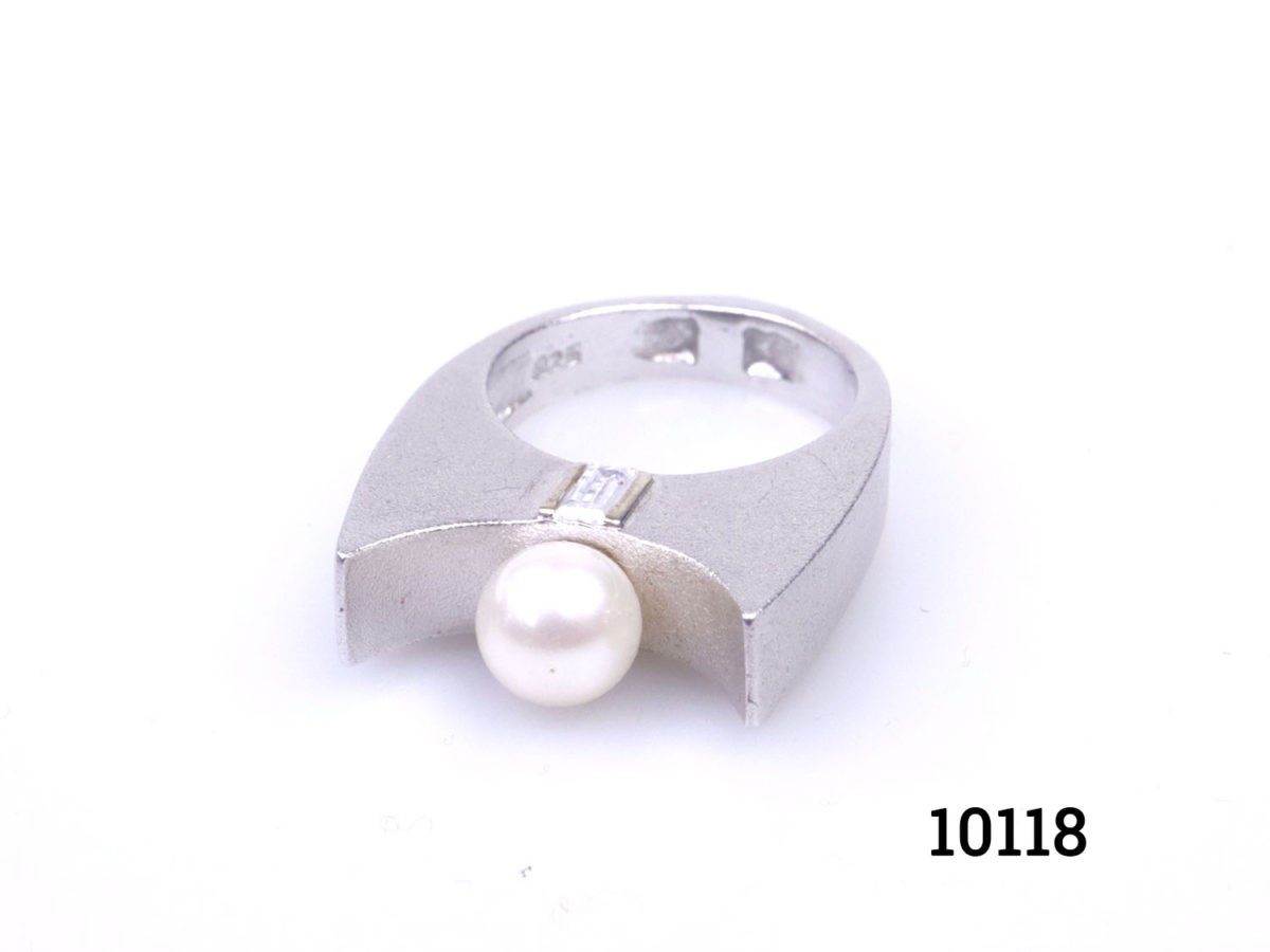 Chunky Modernist style sterling silver ring set with pearl to the centre and cubic zirconia to the sides below the pearl. Ring size P / 7.5. Ring weight 11.9 Photo of ring on a flat surface looking down from a slight raised height showing the cubic zirconia above the pearl abd unusual curved frontage