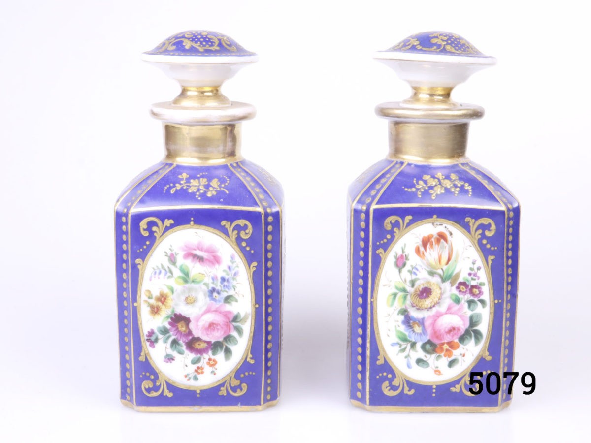 Antique pair of Paris porcelain perfume bottles. Cobalt blue porcelain bottles each decorated with hand-painted floral images and finished in gilt throughout. Some wear to gilt on both bottles. Each bottle measures 75mm by 75mm and 170mm tall. Main photo showing both bottles side by side from an eye level angle with different hand-painted floral decoration visible on each.