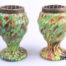 Pair of Art Deco splatter glass posy vases. Some damage to the brass frogs. Each measures 78mm in diameter at base, 90mm in diameter at widest area and 135mm tall.  Main photo of both vases side by side and seen from an eye level angle Photo of