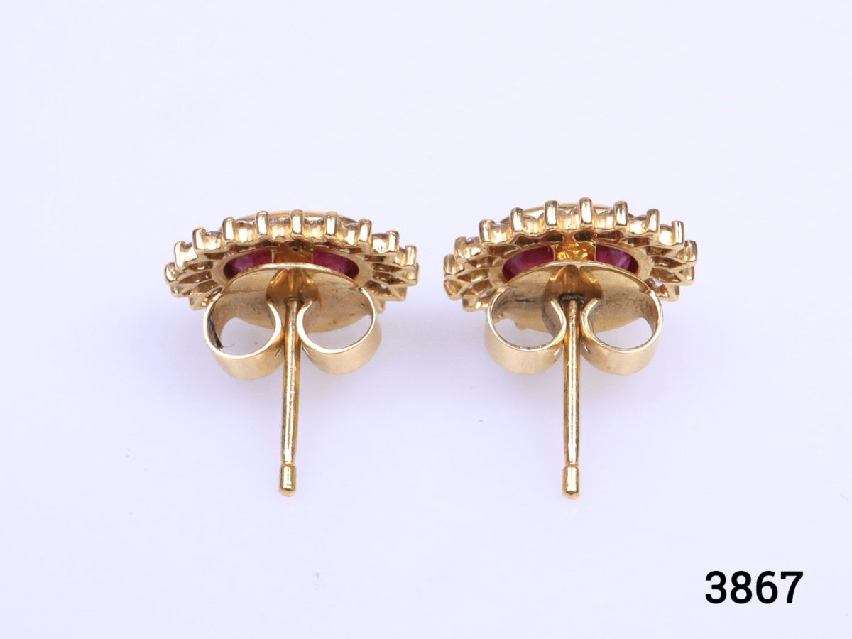 Modern 18 karat yellow gold stud earrings set with oval cut ruby framed in diamonds. Stamped 18k for 18 karat gold. Earrings weigh 5.6 grams and front measures 15mm by 12mm. Box included. Photo of both earrings on a flat surface seen with butterfly fasteners in foreground