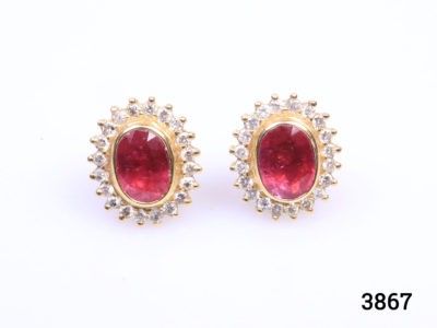 Modern 18 karat yellow gold stud earrings set with oval cut ruby framed in diamonds. Stamped 18k for 18 karat gold. Earrings weigh 5.6 grams and front measures 15mm by 12mm. Box included. Main photo of earrings on a flat surface and seen from the front