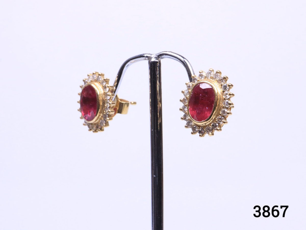 Modern 18 karat yellow gold stud earrings set with oval cut ruby framed in diamonds. Stamped 18k for 18 karat gold. Earrings weigh 5.6 grams and front measures 15mm by 12mm. Box included. Photo of earrings on a display stand and seen from a slight side angle