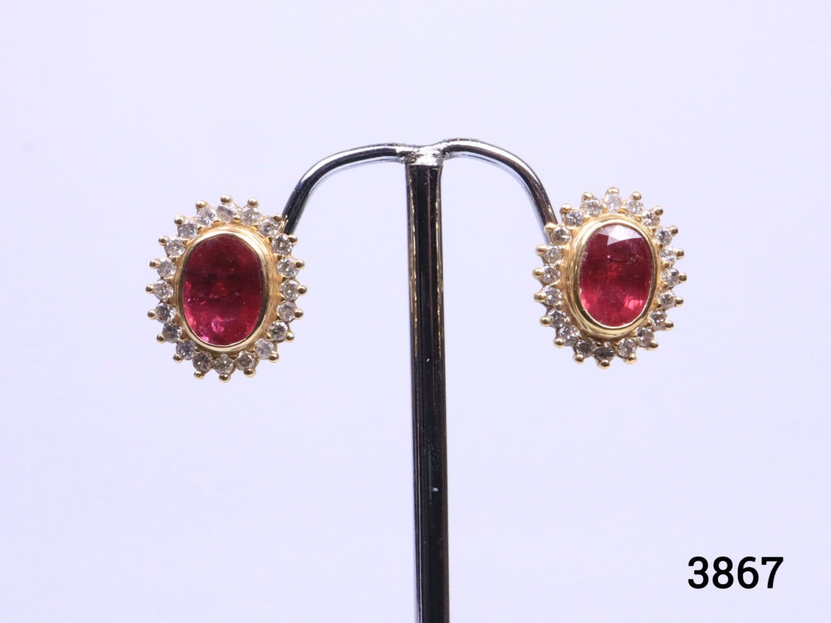 Modern 18 karat yellow gold stud earrings set with oval cut ruby framed in diamonds. Stamped 18k for 18 karat gold. Earrings weigh 5.6 grams and front measures 15mm by 12mm. Box included. Photo of earrings on a display stand and shown from the front