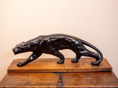Vintage black panther mounted onto a wooden stand. Vintage Art Deco ornament of a black panther mounted onto a wooden stand. Signed to the wooden base.(Damage to from leg where black coating has come away exposing the plaster) Base measures 655mm long by 175mm wide and 30mm tall. Panther measures 570mm long by 200mm high.