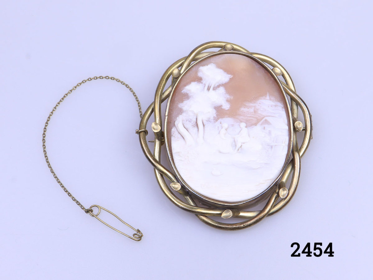Vintage large pinchbeck cameo brooch. Intricately carved cameo of a man and woman holding hands and seated outdoors by a pond. Incredible detailing in the cameo carving. of an unusual scene. Main photo of brooch displayed on a flat surface with safety chain and pin laid out to the left