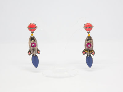 Pair of modern rocket ship costume jewellery earrings. Fun pair of earrings of rocket ships flying towards a pink enamel planet with a black crystal ring. Rocket is made of white metal and shows an alien eye through the porthole and a metallic blue jet flame below. Made in France by Taratata. Main photo of earrings on a clear display stand