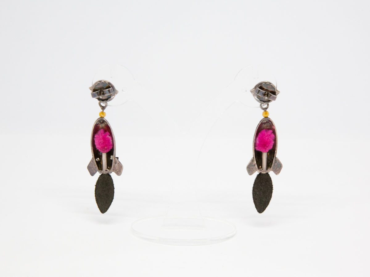 Pair of modern rocket ship costume jewellery earrings. Fun pair of earrings of rocket ships flying towards a pink enamel planet with a black crystal ring. Rocket is made of white metal and shows an alien eye through the porthole and a metallic blue jet flame below. Made in France by Taratata. Photo of earrings on a display stand and seen from the back