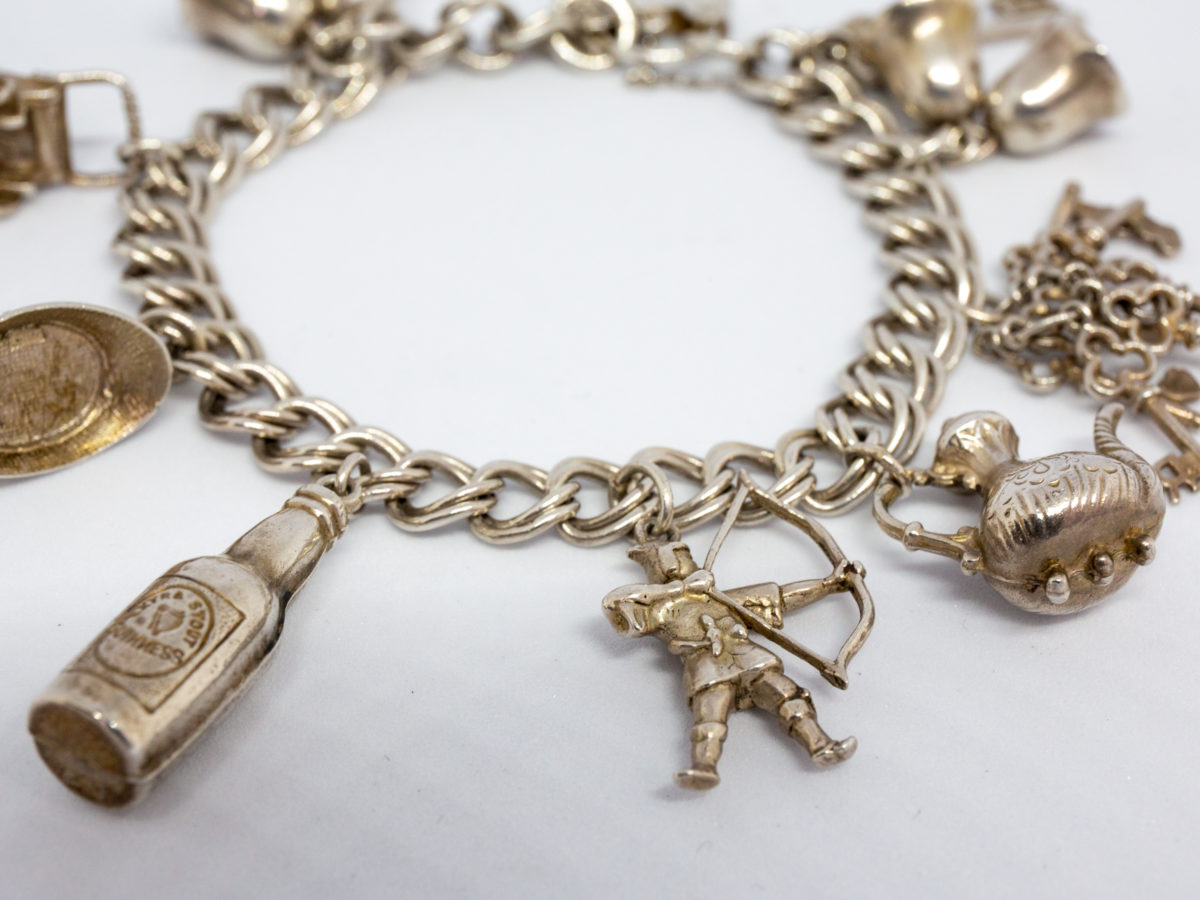 Vintage sterling silver charm bracelet. Double link chain bracelet with lion passant hallmark to most of the chain links and a mixed assortment of 9 charms some with hallmarks. Heart shaped padlock clasp. Bracelet opens to 100mm at widest (safety chain) and closes to approximately 80mm in width. Close up photo of charms (archer, bottle)