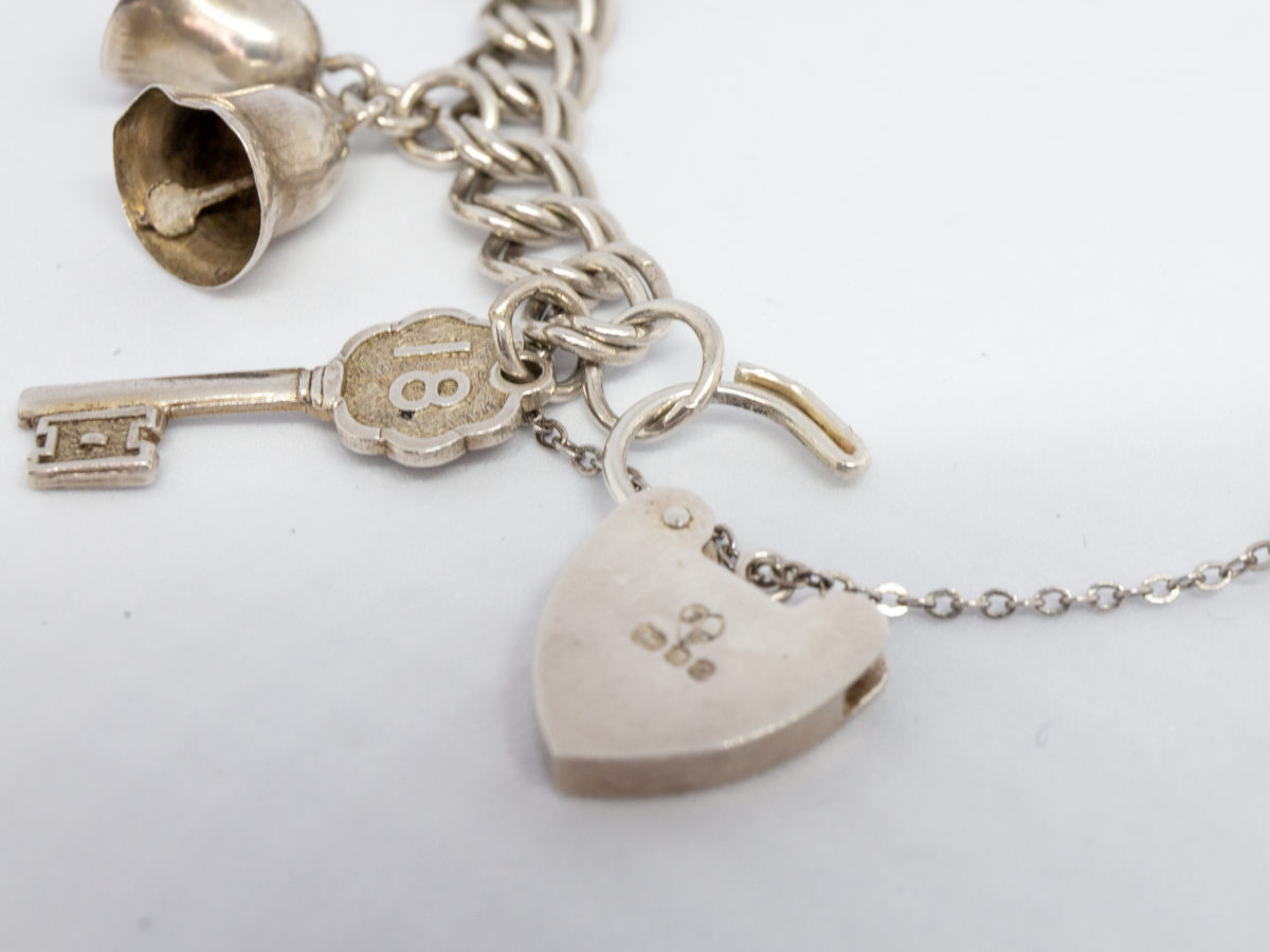 Vintage sterling silver charm bracelet. Double link chain bracelet with lion passant hallmark to most of the chain links and a mixed assortment of 9 charms some with hallmarks. Heart shaped padlock clasp. Bracelet opens to 100mm at widest (safety chain) and closes to approximately 80mm in width. Close up of the heart clasp with catch open