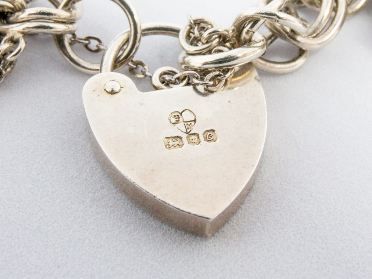 Vintage sterling silver charm bracelet. Double link chain bracelet with lion passant hallmark to most of the chain links and a mixed assortment of 9 charms some with hallmarks. Heart shaped padlock clasp. Bracelet opens to 100mm at widest (safety chain) and closes to approximately 80mm in width. Close up photo of the hallmark on the back of heart clasp