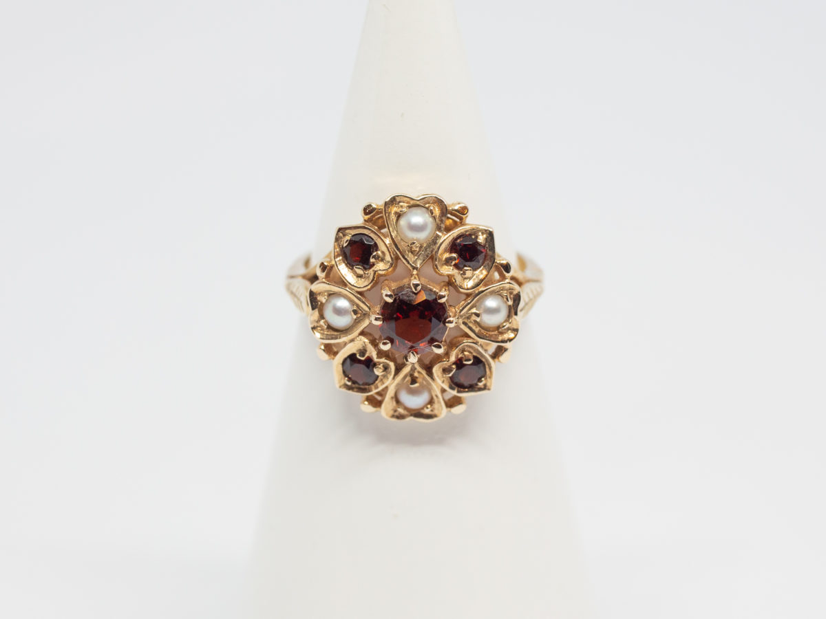 Vintage 9 karat gold ring with garnets and seed pearls. c1959 London assayed 9 karat gold ring with round cut garnet to the centre surrounded by 4 small garnets and seed pearls set on heart shaped petals with leaves on shoulders. Ring size O.5 / 7.75 Ring front measures approximately 18mm by 16mm. Main photo of ring on a display stand shown from the front