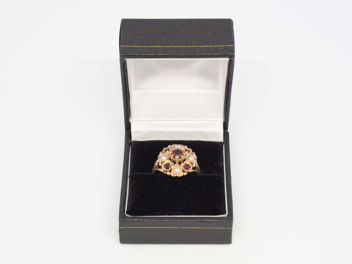 Vintage 9 karat gold ring with garnets and seed pearls. c1959 London assayed 9 karat gold ring with round cut garnet to the centre surrounded by 4 small garnets and seed pearls set on heart shaped petals with leaves on shoulders. Ring size O.5 / 7.75 Ring front measures approximately 18mm by 16mm. Photo of ring displayed in box (included)