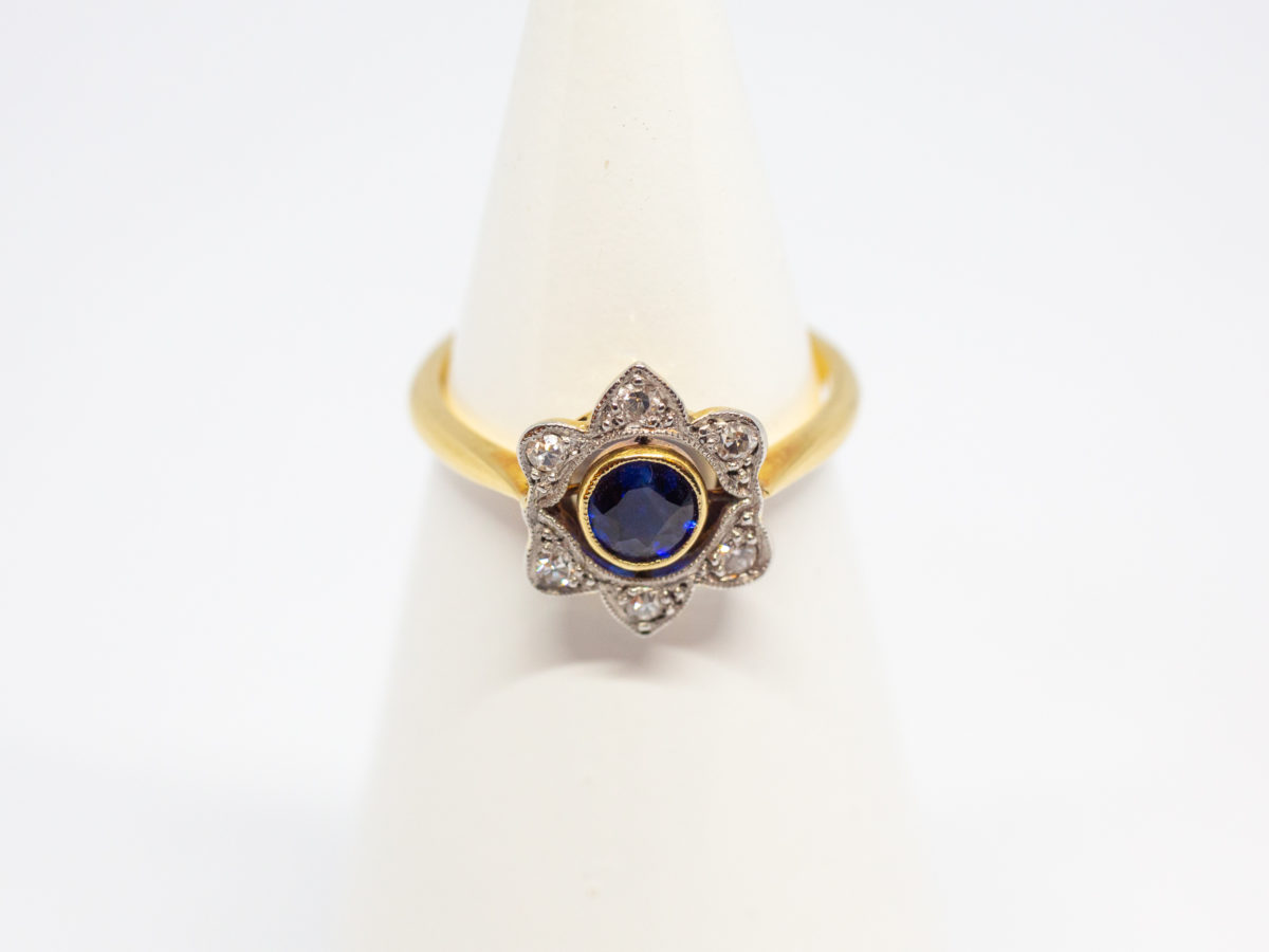 Art Deco sapphire and diamond ring. Pretty ring in the form of a flower with a stunning blue round cut sapphire to the centre with 6 small round cut diamonds on each outer petal. Set on 18 karat yellow gold. Ring size L / 5.75. Weight 2.8gms. Box included. Main photo of ring displayed on a cone stand with ring front to the centre of photo