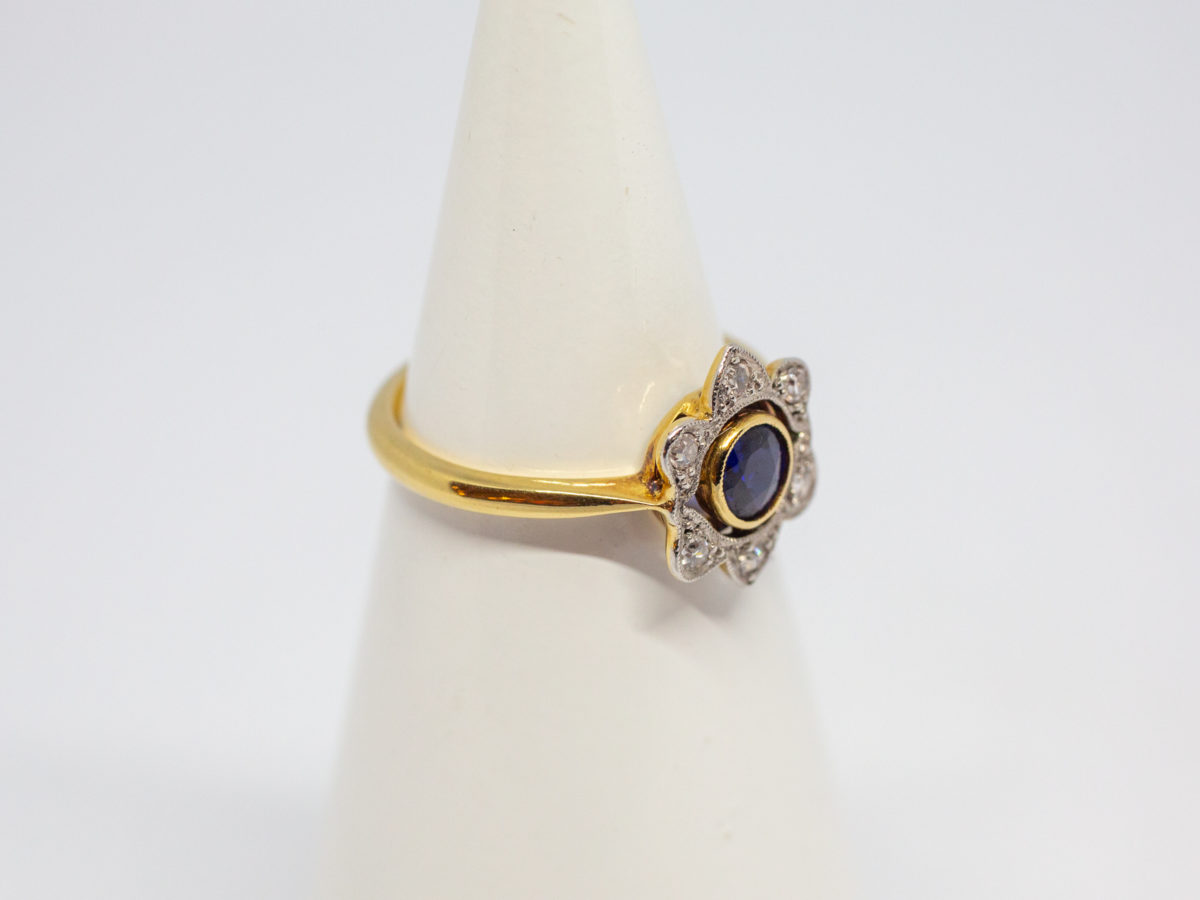 Art Deco sapphire and diamond ring. Pretty ring in the form of a flower with a stunning blue round cut sapphire to the centre with 6 small round cut diamonds on each outer petal. Set on 18 karat yellow gold. Ring size L / 5.75. Weight 2.8gms. Box included. Photo of ring on a cone display stand with ring front facing right.