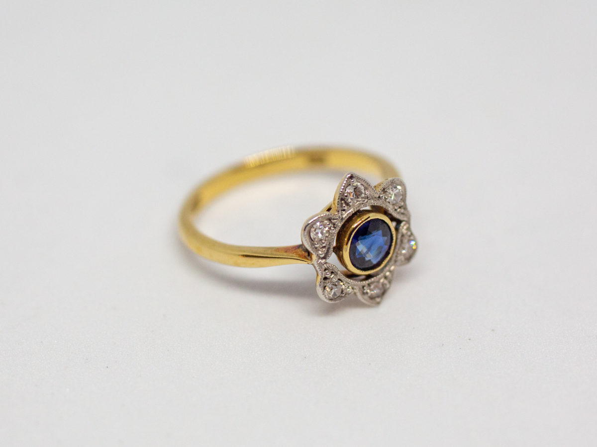 Art Deco sapphire and diamond ring. Pretty ring in the form of a flower with a stunning blue round cut sapphire to the centre with 6 small round cut diamonds on each outer petal. Set on 18 karat yellow gold. Ring size L / 5.75. Weight 2.8gms. Box included. Photo of ring laid on a flat surface and seen with ring front to the bottom right of photo.