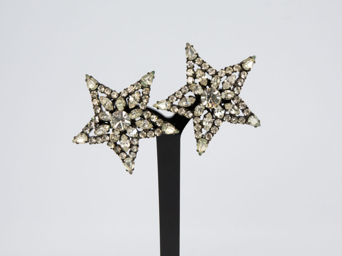 Vintage 1980s Butler & Wilson earrings. Large rhinestone starburst clip-on earrings. Marked B&W to the back. Both clips are in good working order. Each earring measures approximately 50mm in diameter. Main photo of earrings on a display stand and seen from the front (earrings are not shown level as the stand has one arm higher than other)