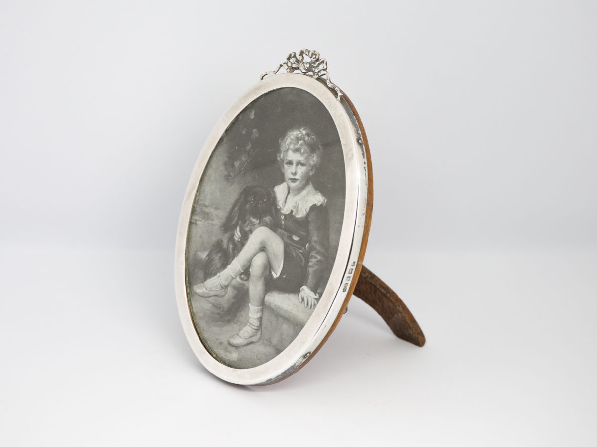 Antique sterling silver oval photo frame. Lovely oval photo frame with pretty ribbon detail to the top. Nice solid wood back. Full hallmark to the side for Birmingham assay c1916. Visible photo area measures 130mm by 88mm at widest. Photo of frame shown upright and from a slight off side angle with the wooden back stand visible to the right of frame front