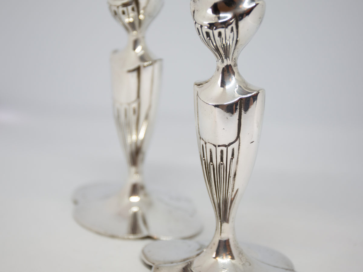 c1920 American Art Deco candlesticks. Stunning pair of shapely Art Deco silver plated candlesticks. Made by Simpson H.M & Co. One candlestick has an uneven base and a slight wobble. Each stick measures approximately 97mm by 75mm at base, 160mm tall at the highest point, 57mm by 48mm across the top and candle slot measures 22mm in diameter. Close up photo of the Art Deco design on the column and capital.