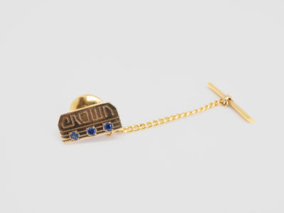 Vintage American 10 karat gold tie tack. Lovely tie tack in 10 karat gold with 3 round cut sapphires below the word crown. (Great sapphire anniversary gift perhaps?) Hallmarked 10k to the back. No hallmark to the pin guard, chain or T-bar. Tack front measures 15mm by 7mm. Main photo of tie tack on a flat surface with chain and T-bar extended out to the right with tack front visible with word 'Crown' and 3 sapphires below.