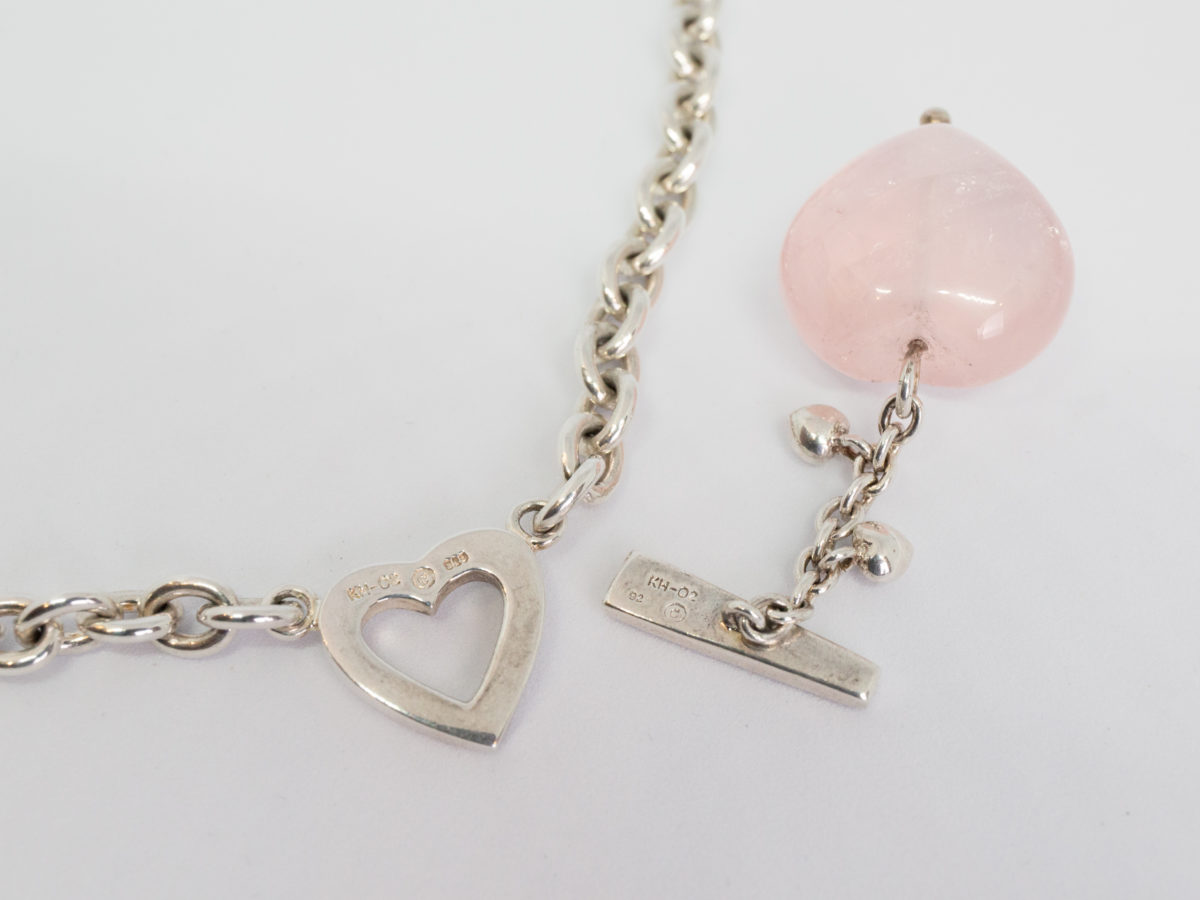 Kit Heath sterling silver and rose quartz necklace. Nice solid silver chain with an attached silver heart and a detachable rose quartz heart. The silver attachment bar is made to resemble an arrow. Rose quartz is believed to attract love and connects to the heart chakra. Detachable heart drop length is 47mm, full drop length of whole pendant area is 53mm. Hallmarked to the clasp, the attached heart and bar of detachable pendant. Close up photo of the hallmarks and also showing the heart section removed from the main necklace.