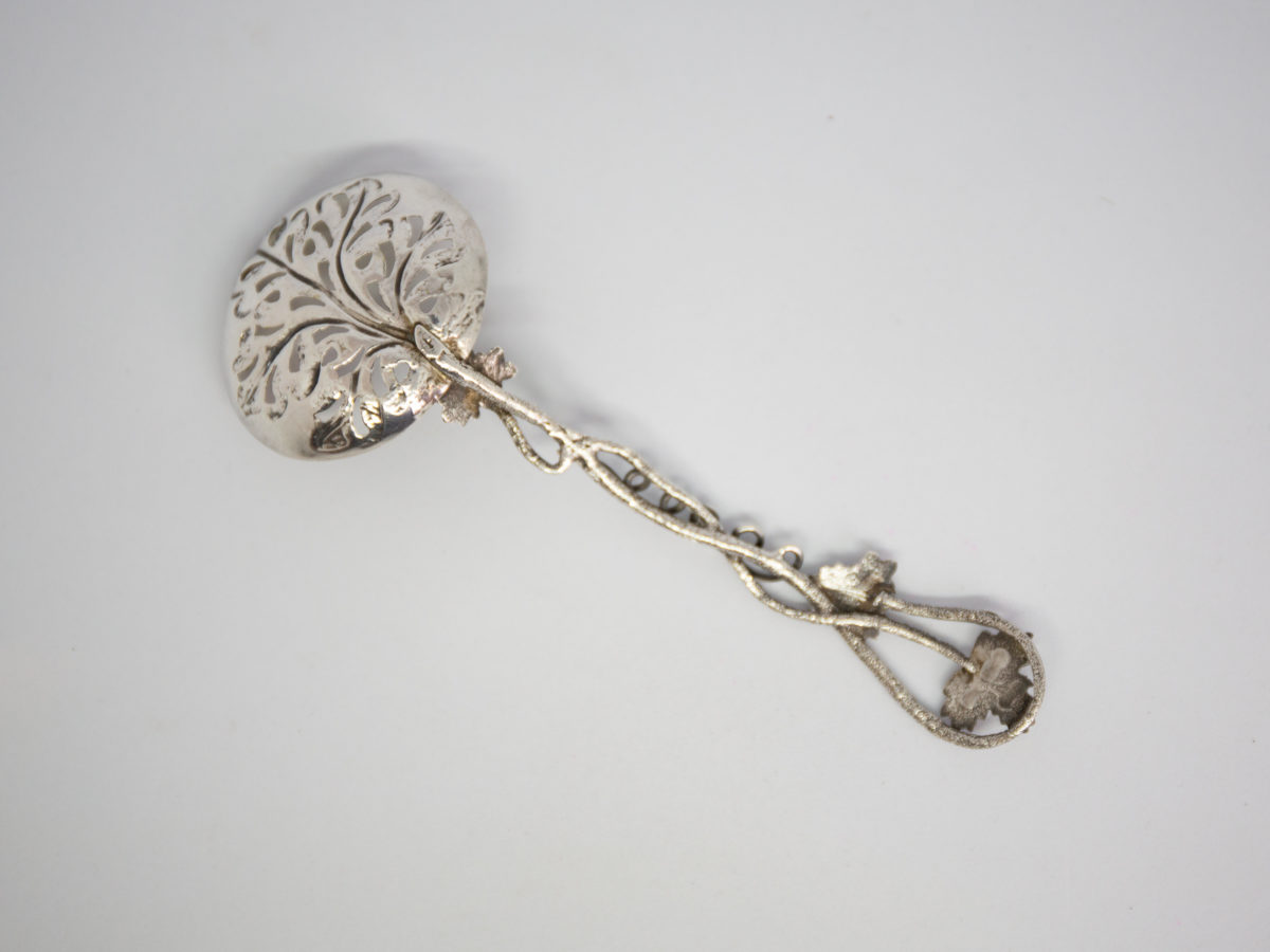c1853 Sterling silver fruit strainer spoon. Very fine quality & beautifully crafted sterling silver fruit strainer spoon in a vine pattern. The handle is made to resemble vine stalks and leaves with an extended vine leaf design for the spoon bowl which is finished in gilt. Full hallmark to the top vine leaf for Birmingham assay and made by Hilliard & Thomason. Spoon bowl measures 45mm by 40mm. Photo of back of spoon with spoon bowl in top left corner of photo. Photo of back of spoon with focus on the spoon bowl which is in the bottom left corner of photo.