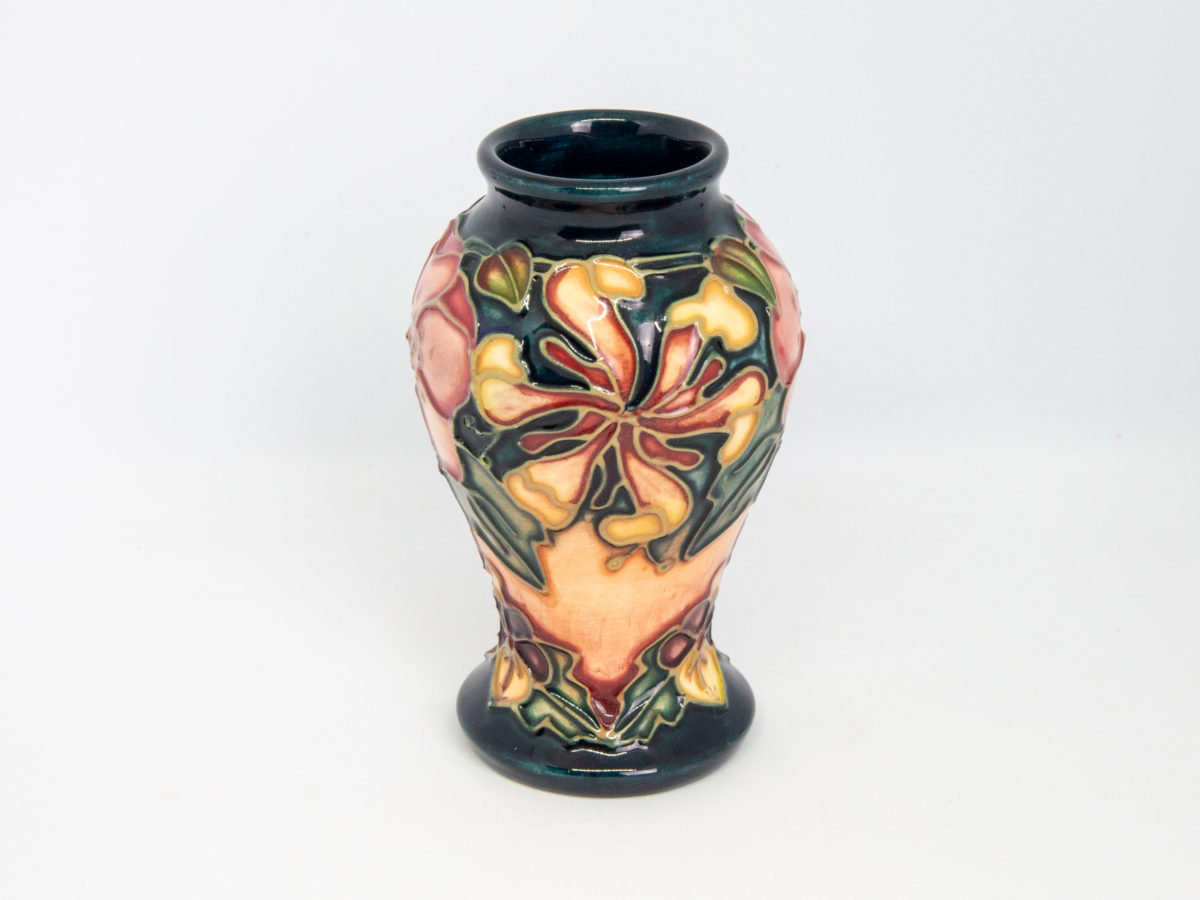 Moorcroft Oberon honeysuckle vase. Small Moorcroft vase in classic Moorcroft colours and decorated in the Oberon honeysuckle pattern. Markings to the base are faded but still just readable. Designed by Rachel Bishop and dated c1993. Measures 46mm in diameter at base, 57mm at widest point and 34mm across the top. Photo of vase showing a side with fully blooming honeysuckle flower.