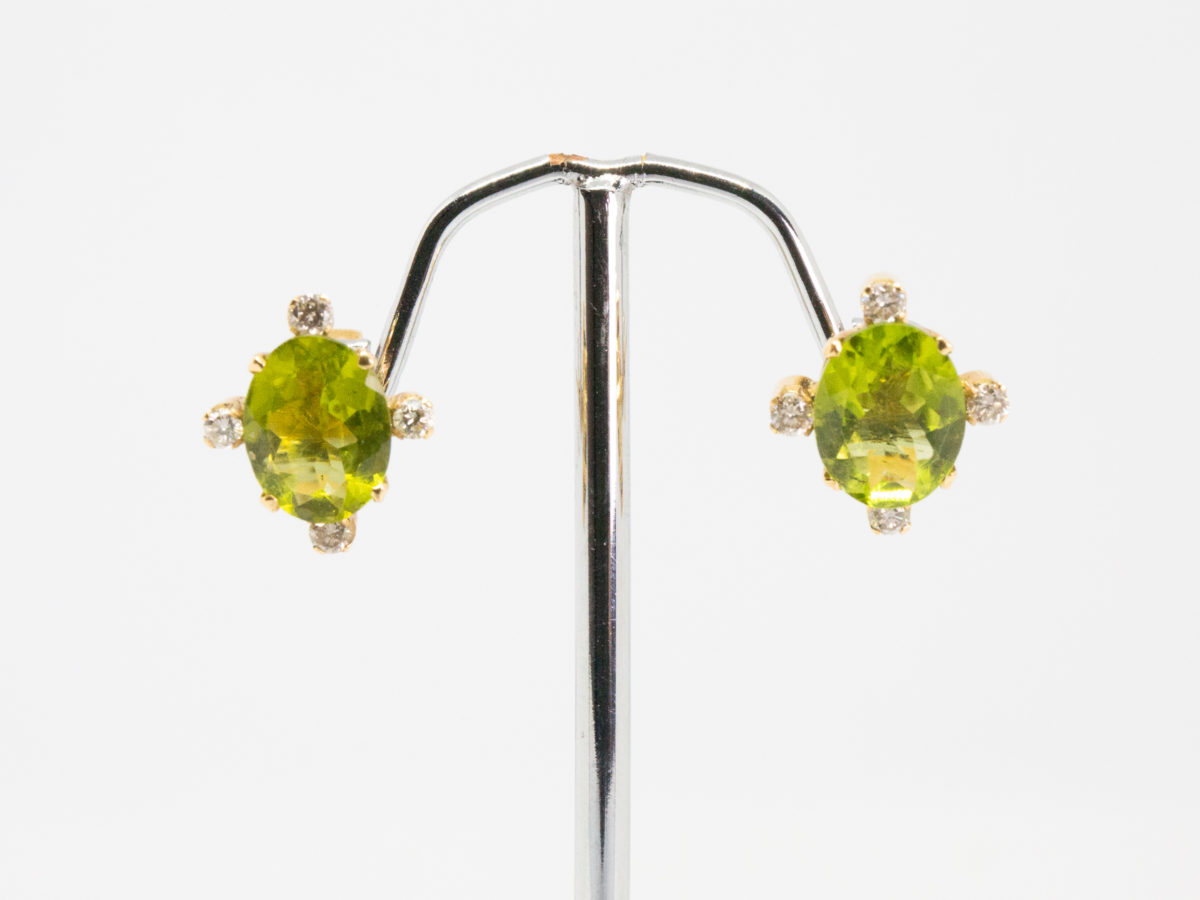 Modern 18 karat gold peridot and diamond earrings. Gorgeous stud earrings set to the centre with an oval cut peridot with a small round cut diamond to each of its 4 sides. Hallmarked 18k to the back of each stud and to each butterfly. Each earring front measures 13mm by 12mm. Box included. (Not shown) Main photo of earrings displayed on a stand with earring fronts facing forward.