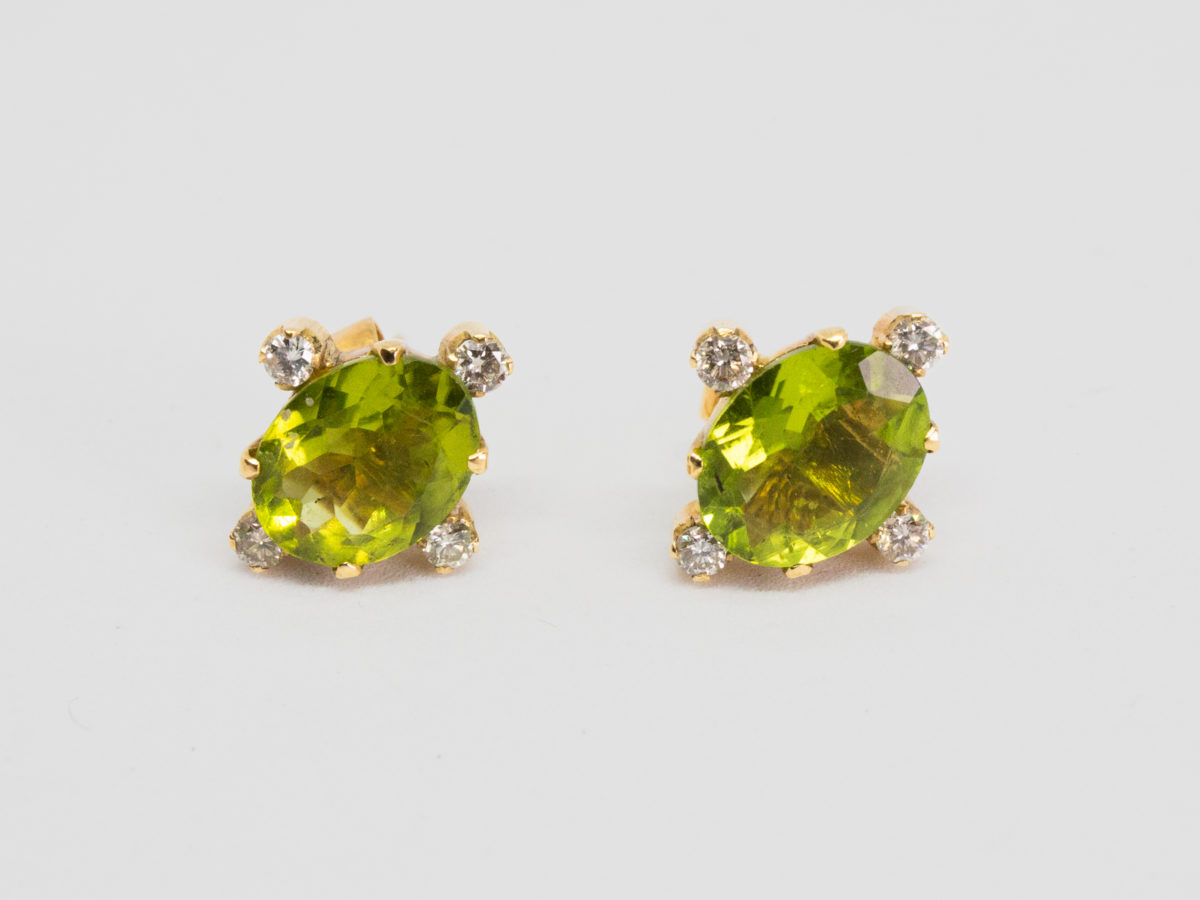 Modern 18 karat gold peridot and diamond earrings. Gorgeous stud earrings set to the centre with an oval cut peridot with a small round cut diamond to each of its 4 sides. Hallmarked 18k to the back of each stud and to each butterfly. Each earring front measures 13mm by 12mm. Box included. (Not shown) Photo of both earrings laid on a flat surface set at a slight diagonal to accommodate a diamond at the tip.