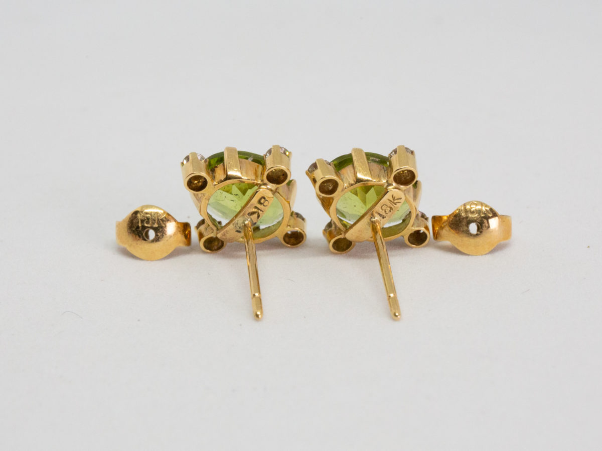 Modern 18 karat gold peridot and diamond earrings. Gorgeous stud earrings set to the centre with an oval cut peridot with a small round cut diamond to each of its 4 sides. Hallmarked 18k to the back of each stud and to each butterfly. Each earring front measures 13mm by 12mm. Box included. (Not shown) Photo of back of both earrings with butterfly fasteners removed and showing the 18kt hallmark to the back of each earring and butterfly.