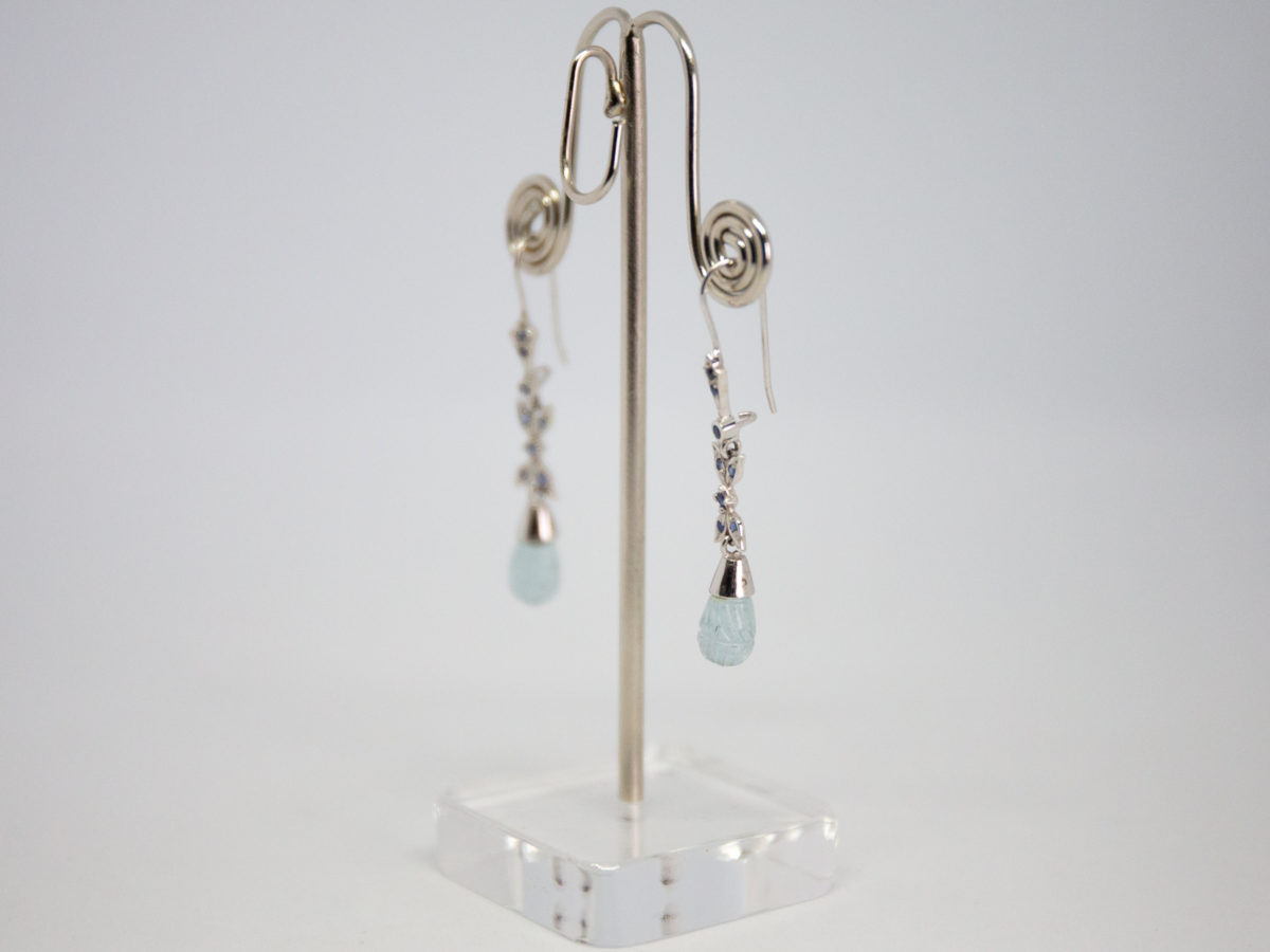 Modern 18 karat white gold, aquamarine and sapphire earrings. Sophisticated & sweet dangle earrings in 18 karat white gold set with small round cut sapphires and a droplet of carved aquamarine to finish off at the ends. Earring drop length approximately 50mm from top of hook to bottom of aquamarine droplet. Phot of earrings displayed on a stand at a diagonal angle with earring on the right closer to foreground.