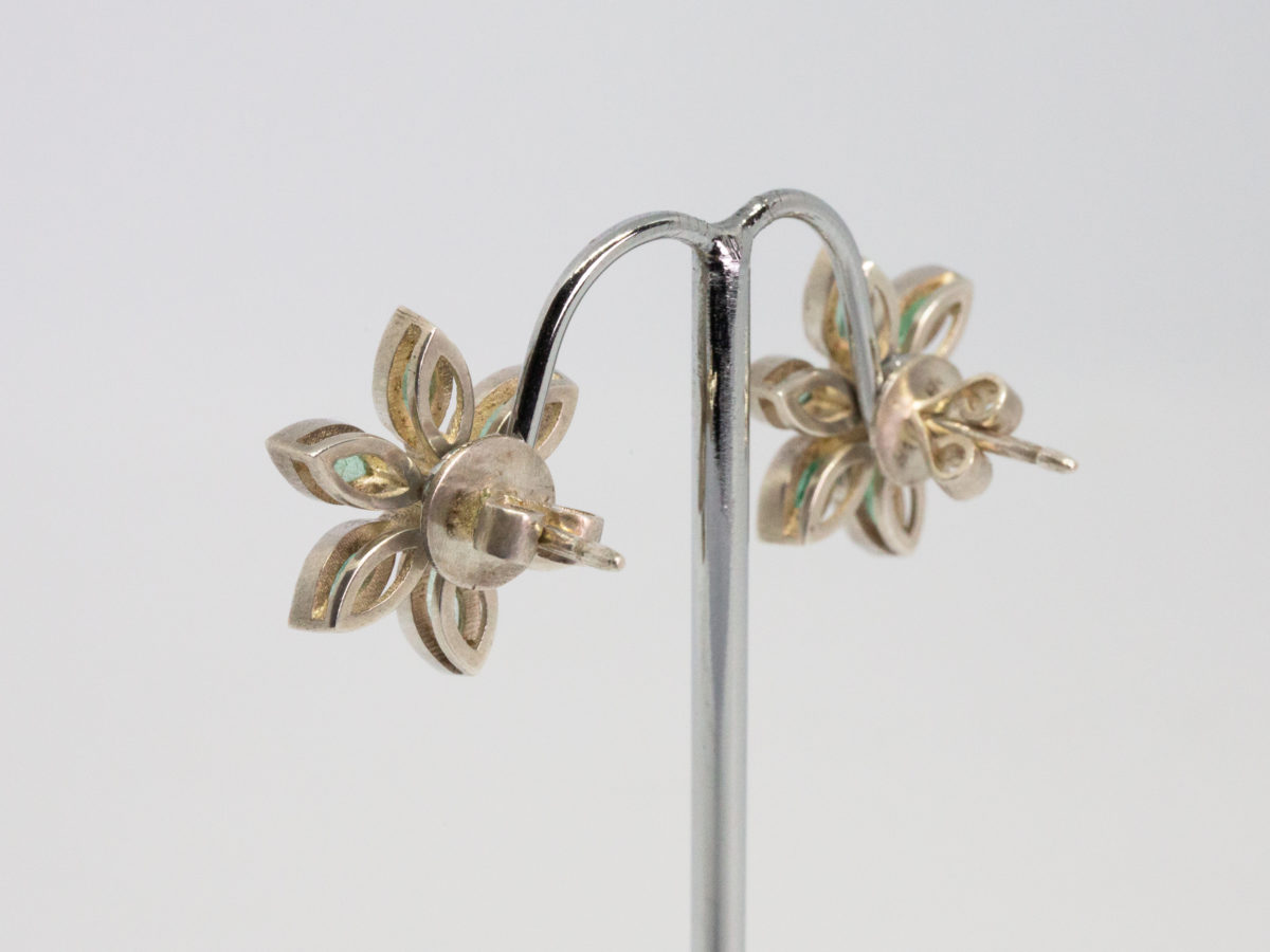 Modern sterling silver, emeralds and sapphire stud earrings. Very pretty pair or sterling silver stud earrings in a floral design with oval cut emeralds as the flower petals and a single round cut white sapphire to the centre. Earring front measures approximately 20mm in diameter. Photo of both earrings displayed on a stand and showing the back with butterflies attached.
