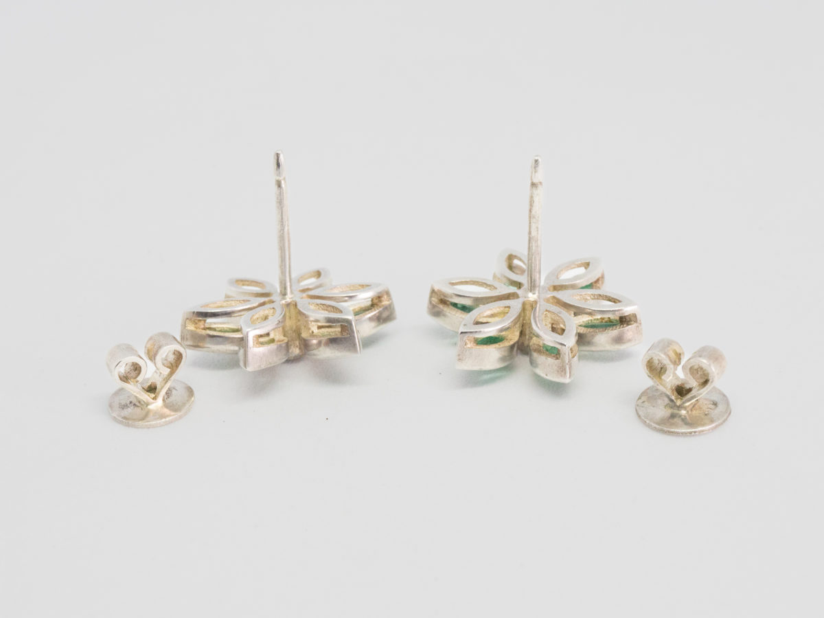 Modern sterling silver, emeralds and sapphire stud earrings. Very pretty pair or sterling silver stud earrings in a floral design with oval cut emeralds as the flower petals and a single round cut white sapphire to the centre. Earring front measures approximately 20mm in diameter. Photo of both earrings face down on a flat surface with the butterfly fasteners removed.