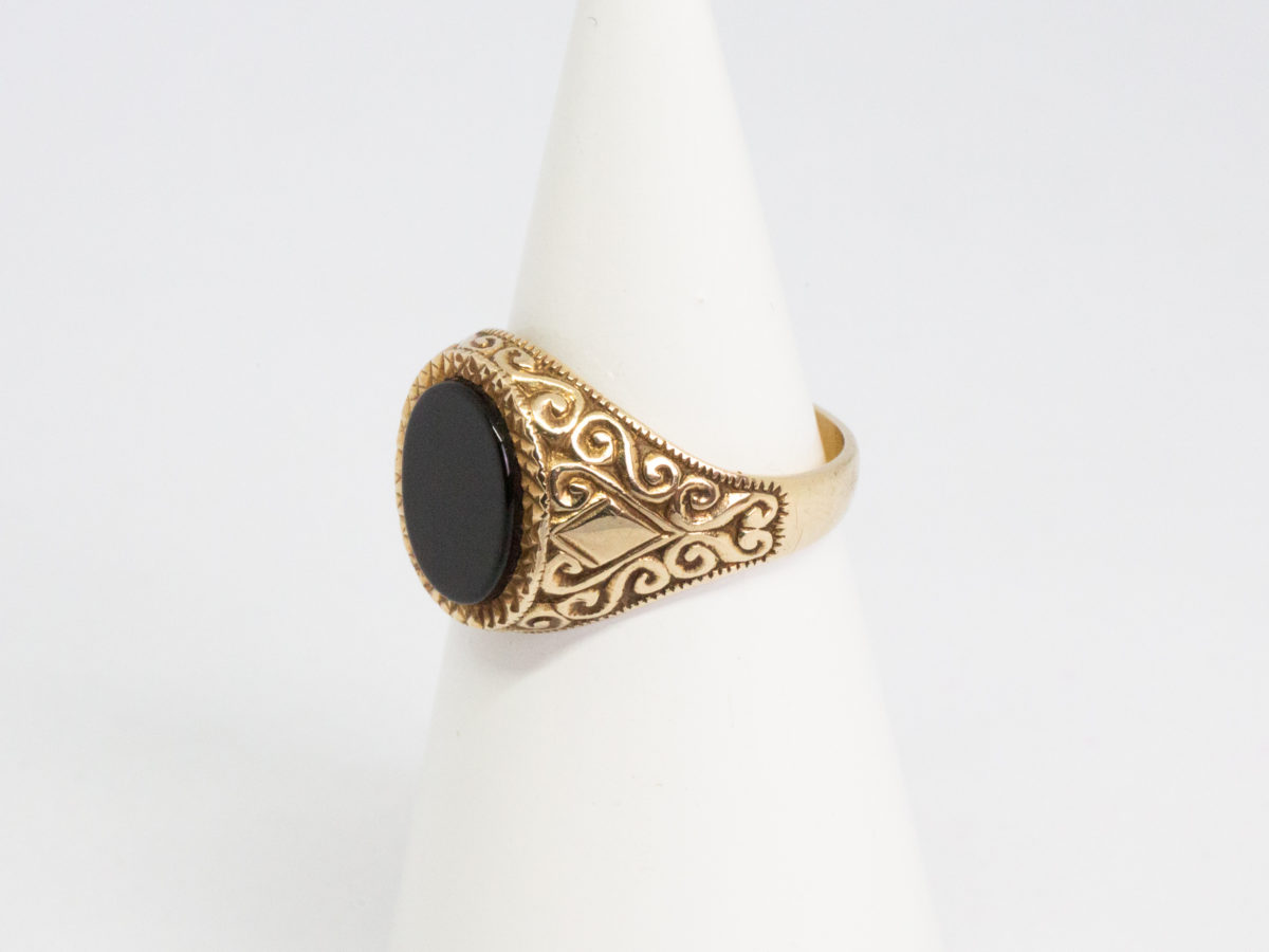 Vintage 9 karat gold and black onyx ring. Smooth oval black onyx to the centre of fine cut mini pyramid frame with fancy scrollwork to each shoulder. Fully hallmarked for Continental import and assayed in Birmingham. Ring size R.5 / 9. Photo of ring displayed on a cone shaped stand with ring front facing left of photo and showing the scrollwork detail on the shoulder.