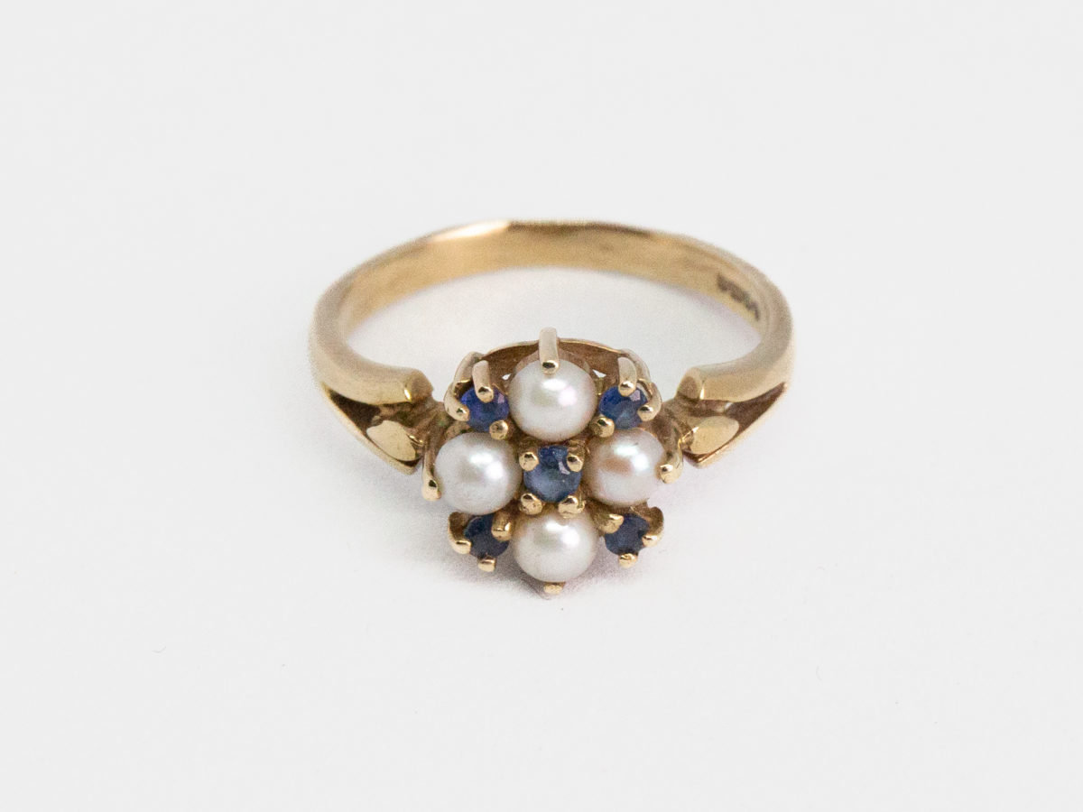 Vintage 9 karat gold, sapphire and pearl ring. Pretty 9 karat gold ring set with 5 small round cut blue sapphires and seed pearls. Hallmarked for Continental import with Birmingham assay. Ring size Q / 8. Ring weight 3.2gms. Photo of ring on a flat surface with ring front in foreground. The hallmark can just be seen to the right of inside band.