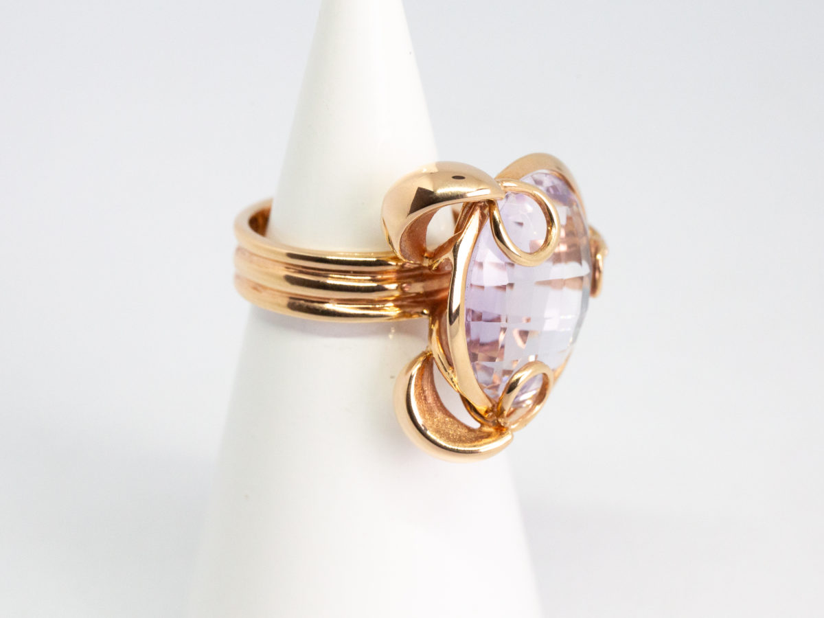 18 karat rose gold and amethyst ring. A stunning statement ring in 18 karat rose gold with a large faceted round cut lilac amethyst stone. Unusual raised setting with 3 hoop claws to secure the stone. Looks incredible worn! Italian made quality design. Hallmarked to the inside band. Ring size M / 6. Weight 12.7gms. Photo of ring on a cone shaped display stand and seen with ring front facing right.