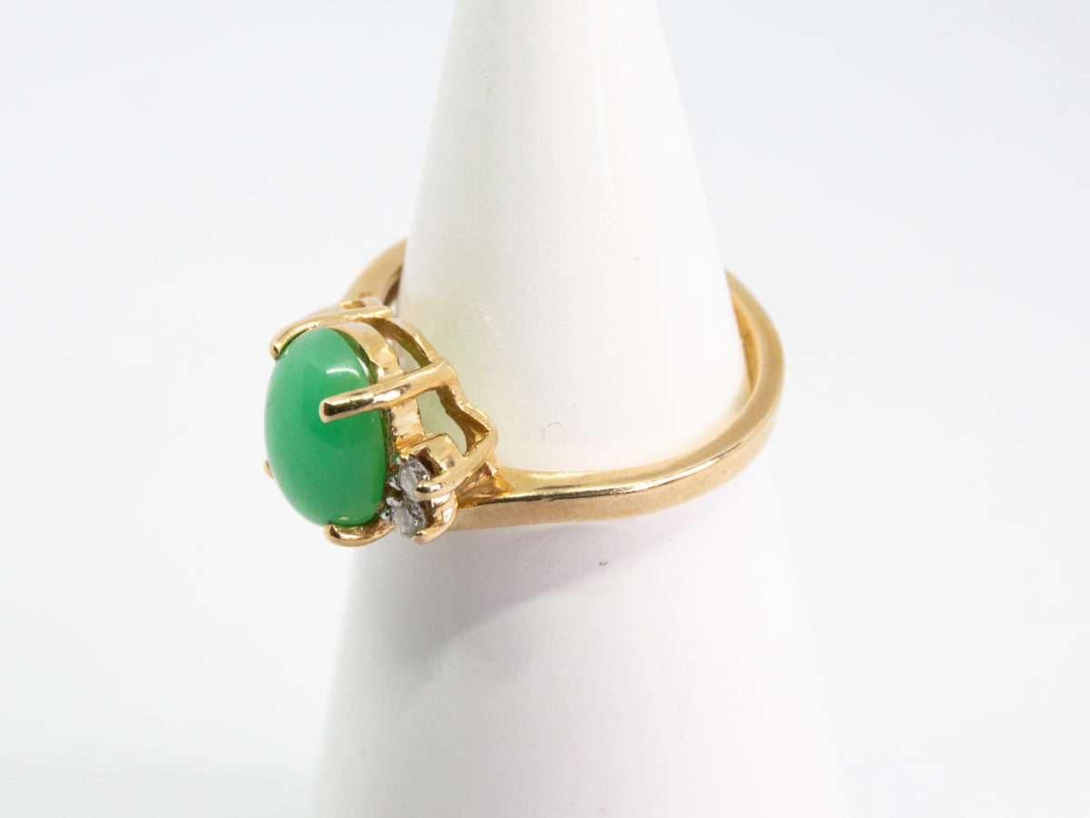 9 karat gold jade and diamond ring. Pretty 9 karat gold ring set with an apple green jade to the centre with 2 small round cut diamonds to one side & 2 gold wave curls to the other side. Hallmarked 375 to inside band. Ring size M.5 / 6.5. Weight 3.2gm. Photo of ring on a cone display stand and shown with ring front facing left.