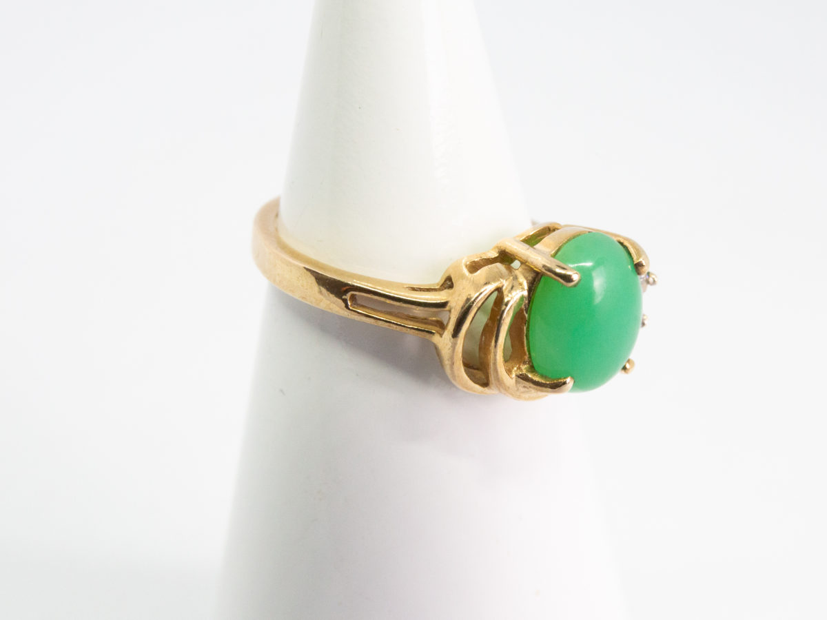 9 karat gold jade and diamond ring. Pretty 9 karat gold ring set with an apple green jade to the centre with 2 small round cut diamonds to one side & 2 gold wave curls to the other side. Hallmarked 375 to inside band. Ring size M.5 / 6.5. Weight 3.2gm. Photo of ring displayed on a cone shaped stand with ring front facing right.