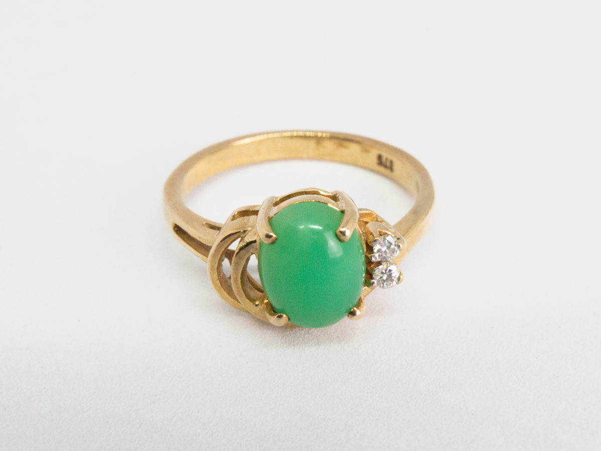 9 karat gold jade and diamond ring. Pretty 9 karat gold ring set with an apple green jade to the centre with 2 small round cut diamonds to one side & 2 gold wave curls to the other side. Hallmarked 375 to inside band. Ring size M.5 / 6.5. Weight 3.2gm. Photo of ring on a flat surface and front facing with diamonds to the right and gold waves to the left.