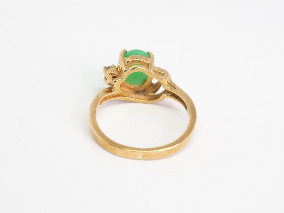 9 karat gold jade and diamond ring. Pretty 9 karat gold ring set with an apple green jade to the centre with 2 small round cut diamonds to one side & 2 gold wave curls to the other side. Hallmarked 375 to inside band. Ring size M.5 / 6.5. Weight 3.2gm. Photo of ring seen from the back with back of band in foreground.
